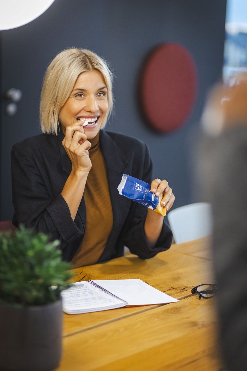 A smiling blonde woman in a dark blazer sits at a wooden table, eating a LIVAGLUCO® snack bar from its blue wrapper. A notebook, pen, and plant are also on the table.