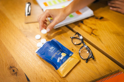 A hand reaches for white tablets near a blue and yellow LIVAGLUCO® sachet on a wooden table, alongside eyeglasses, a notebook, and papers.